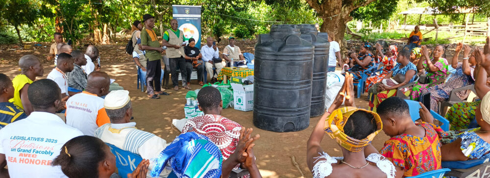 Community members gathered outdoors under large trees during a training and distribution event, seated in a circle around water storage tanks and agricultural supplies while speakers address the group.