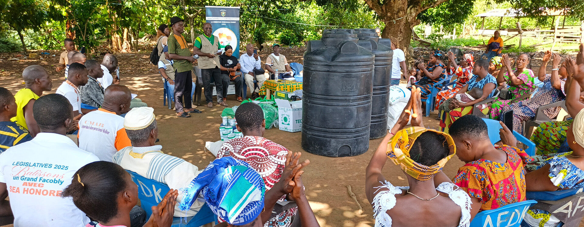 Des membres de la communauté sont réunis en plein air sous de grands arbres lors d’une séance de sensibilisation et de distribution de matériel. Ils sont assis en cercle autour de réservoirs d’eau et de fournitures agricoles tandis que des intervenants s’adressent au groupe.
